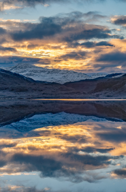Still water reflects sunrise and snowy landscape.