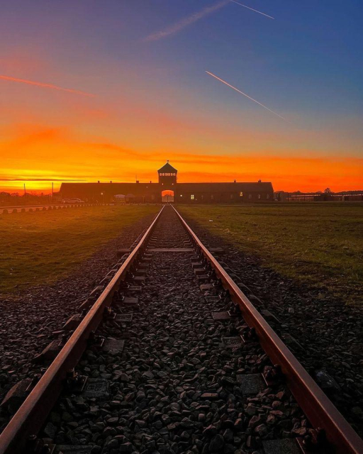 The iconic railway tracks leading into the Auschwitz-Birkenau concentration camp, framed under a vibrant orange and blue sunset sky. The tracks converge at the main entrance gate with the guard tower silhouetted against the fiery hues of the setting sun.
