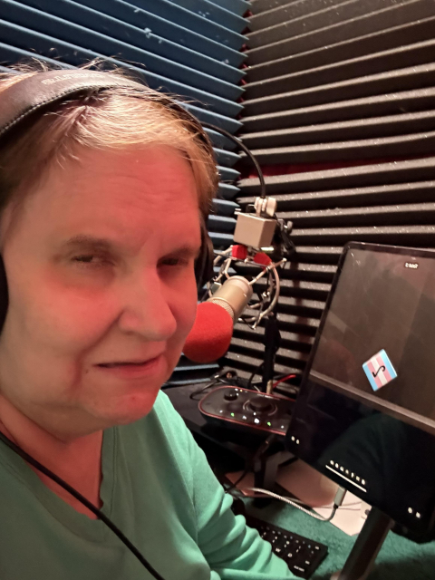 Shelly sits in her small podcast studio. A microphone and an iPad with a dice rolling app visible can be seen.