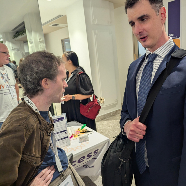 Michael Curran in a brown jacket talks with Michael Sheppard in a blue suit & tie as James Boreham in NV Access shirt talks with another attendee in the background.