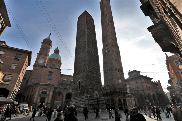 Photo of the famous square in the center of Bologna where two square stone towers on opposite sides of a street stick way, way, way, way up above the other buildings. Their bare stone sides are pierced only by a few very small windows, and undecorated. They are wrapped in modern metal bracing to help protect them from crumbling.