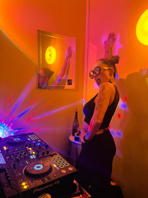 a femme dj in the booth, wearing all black, orange and bright colored lights, sexy and silly