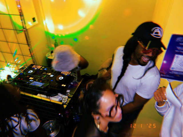 a black and queer dj collective in the booth. the femme with the hat is on the decks, the man is standing and smiling, i'm moving too fast and am blurry. 