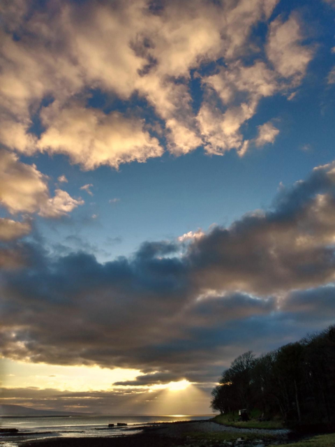 In front and to the right, the shore and the forest of Lissadell lie almost in the dark. Further to the left and behind, the ocean, shimmering in the evening light, stretches as far as the horizon. Above it is a wide sky with the light of the setting sun shining through the clouds.