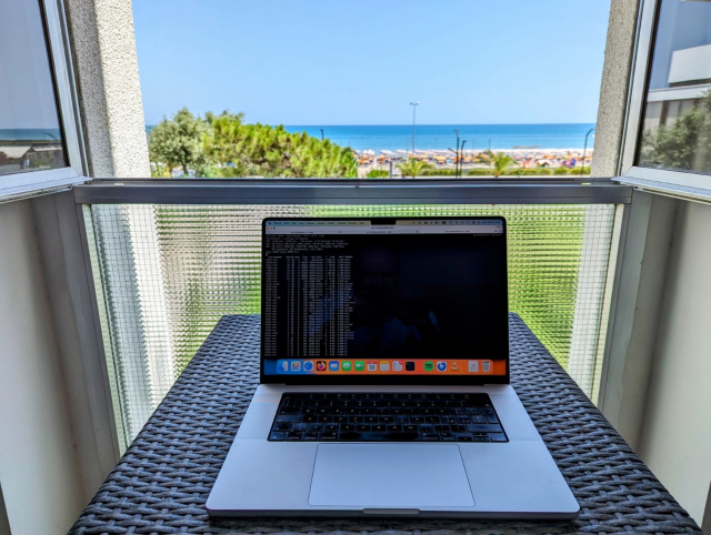 A laptop placed on a woven table next to a window overlooking a beach scene. The laptop screen displays a dark interface with lines of text. Outside, the view reveals a clear blue sky, palm trees, and a sandy beach with sunbathers in the distance. The window has white frames and a partially pulled-down mesh screen.