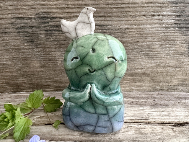 against a background of grey-brown driftwood, a raku ceramic earth spirit jizo sculpture. they have a gentle, smiling face and two little praying hands. there's a white bird on top of their head (it's their friend). next to them on the left are some green speedwell leave and a blue speedwell flower.