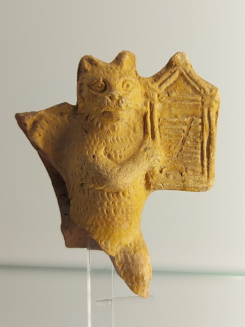 The picture shows a terracotta figure of a standing cat playing a harp-like instrument with its front legs. The lower part of the figurine is broken off.