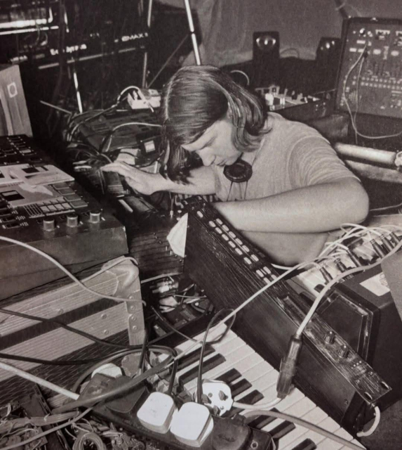 Black and white photograph of a young Richard D James, aka Aphex Twin, in a tiny room surrounded on all sides by synths, drum machines, effects and guitar pedals, like a baby Techno chick in its nest.

There are chaotic strings of cables and power leads strewn about, in the foreground is a 4 gang extension lead, fully utilised with other plugs laying beside it, rested haphazardly on a synth keyboard.

© Photographer unknown