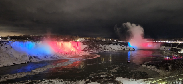 Photo of Niagara Falls taken from the Canadian side at night. Blue, white, and red lights illuminate the misty falls. The sky is clouded with dark grey skies. Snow hugs the edges of the escarpement. City lights are visible in the distance. Ice has collected on top of the water.