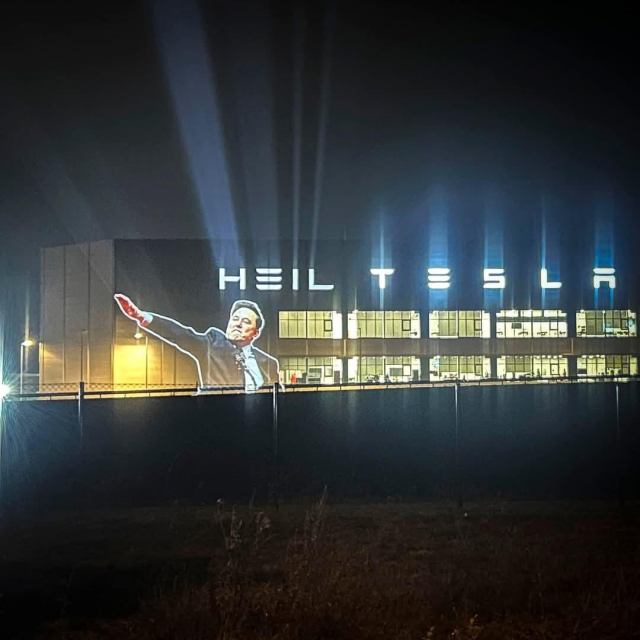 A nighttime projection on the Tesla Gigafactory in Berlin, Germany, displays an image of Elon Musk performing a Nazi-style salute, accompanied by the illuminated text "HEIL TESLA" in Tesla's signature font. This provocative display critiques Musk's recent gesture during President Donald Trump's inauguration, which many interpreted as a fascist salute. The incident has sparked widespread debate and condemnation, with some defending Musk's action as an awkward expression of enthusiasm, while others view it as a dangerous endorsement of far-right ideologies.