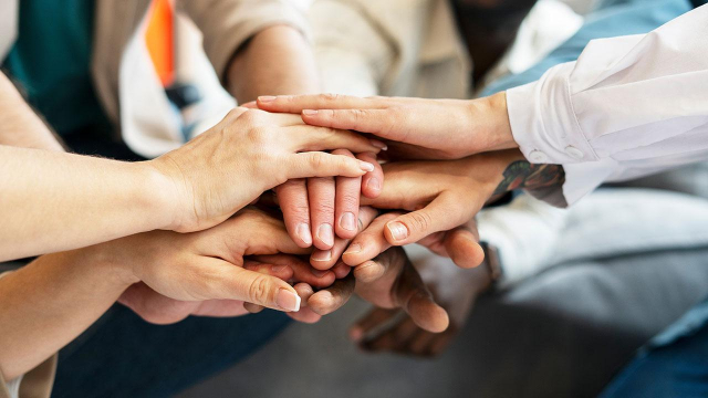 A close-up photograph of multiple hands stacked together in a gesture of unity and teamwork. The hands belong to a diverse group of individuals, showcasing different skin tones, textures, and some adorned with tattoos. The background is blurred, with hints of casual and business attire visible, suggesting a professional or collaborative setting. The image conveys a strong sense of solidarity, cooperation, and mutual support.
