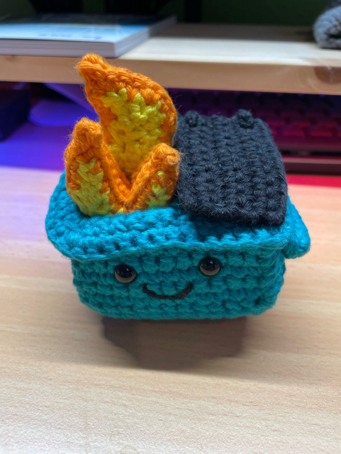 Teal crochet dumpster fire amigurumi on a wooden table