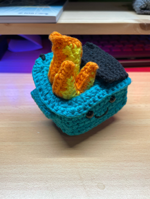 Teal crochet dumpster fire amigurumi on a wooden table