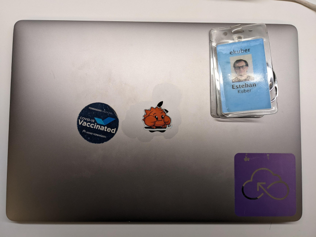 A photo of a MacBook closed on a table with a blue badge on top. Covering the apple logo, a sticker of a cartoonish red crab in a cute pose.