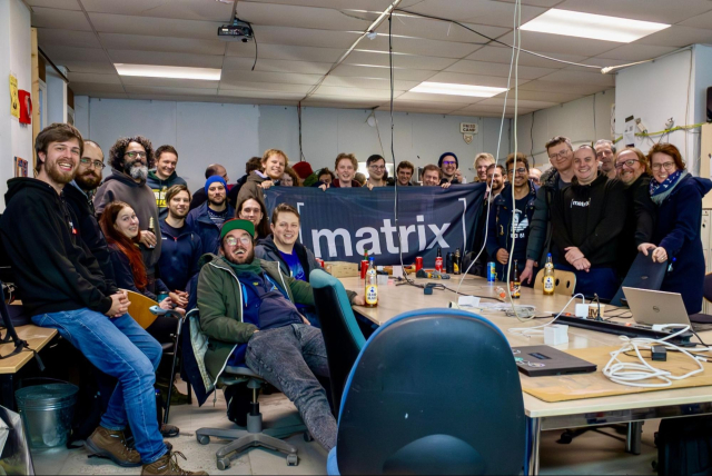 A picture of 30 happy people in a messy room. Thee are cables dangling from the roof and a large Matrix flag. Everyone is smiling or laughing.