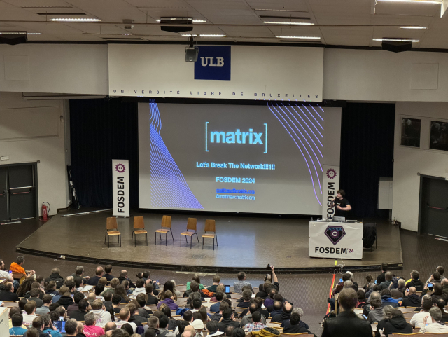 A picture of Matthew Hodgson on the stage of a large amphitheater during FOSDEM 2024, in front of a presentation displaying the Matrix logo prominently. The room is full, there is not a single free seat.