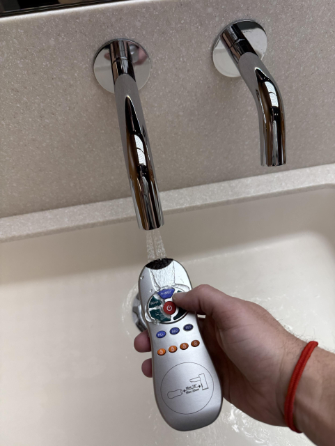 A hand holding a remote control is activating a faucet, with water flowing into a sink below. The faucet has a sleek, modern design. The remote is being soaked.