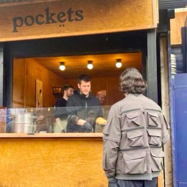 A person stands, back to the camera, in a jacket with 6 large pockets on the back of his jacket. In front of them is a server behind a concession stand labeled “pockets.”