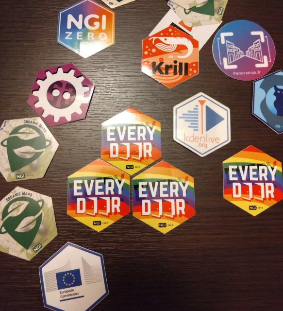 Bunch of hexagonal stickers from NLNet. In the center — Every Door with doors for O's, all in rainbow colors. Also visible Organic Maps, FOSDEM, Kdenlive, Panoramax, Krill, and NGI Zero.