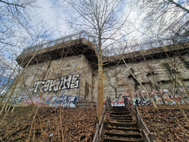 Ansicht eines ehemaligen Flakturms im Volkspark Humboldthain in Berlin-Wedding. Die Betonmauer ist mit Graffiti bedeckt, davor führt eine verwitterte Treppe durch kahle Bäume.

