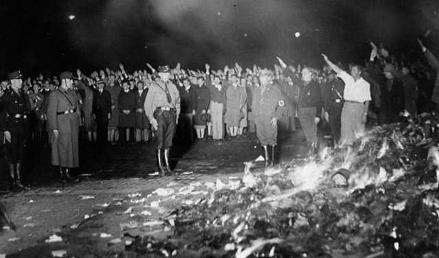 A night shot of crowds looking on and giving Hitler salutes as a bonfire of books and paperwork burns in the foreground. Several figures in the middle ground are wearing Nazi uniforms or have swastika armbands