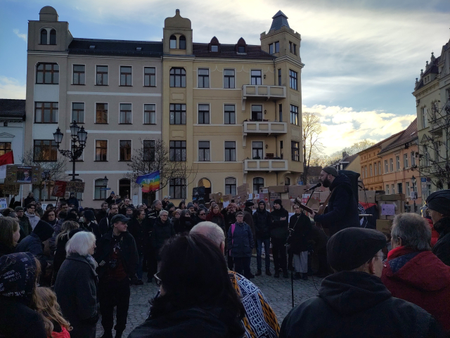 Gruppe von Menschen bei der demo. Einrregenbogenflagge ist zu sehen und im Hintergrund eine aus Kartons aufgebaute Brandmauer.
