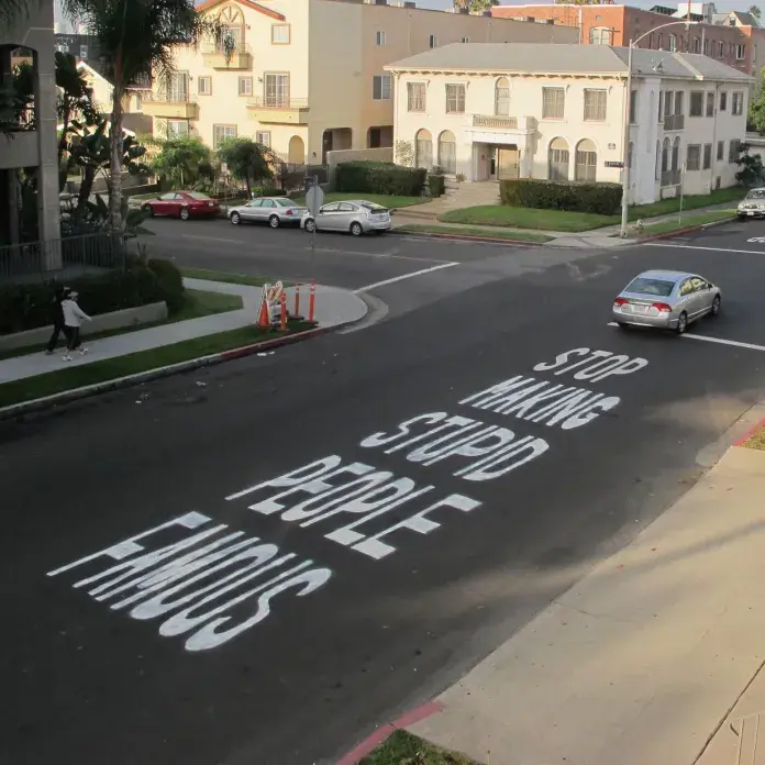  Large white capital letters painted directly onto an asphalt street in Los Angeles spell out the bold statement: "STOP MAKING STUPID PEOPLE FAMOUS." The message, attributed to street artist Plastic Jesus, takes up a significant portion of the road, making it highly visible to passing cars and pedestrians. The urban setting features residential buildings, parked cars, and a person walking on the sidewalk. The artwork critiques modern celebrity culture and the media's role in elevating individuals without merit.
