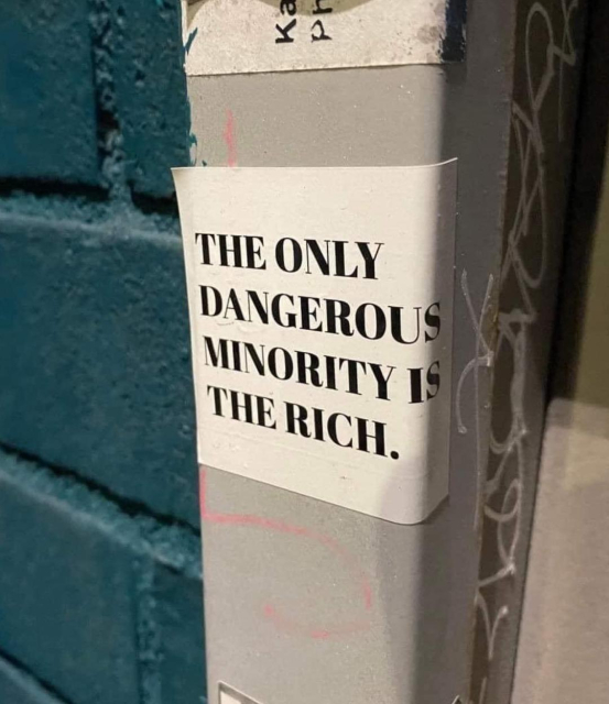 A bold black-and-white sticker with capitalized text reading, "THE ONLY DANGEROUS MINORITY IS THE RICH." The sticker is placed on a metal pole covered in graffiti and scratches, positioned next to a textured blue brick wall. The statement is a striking political message critiquing wealth inequality, blending seamlessly into the urban environment.