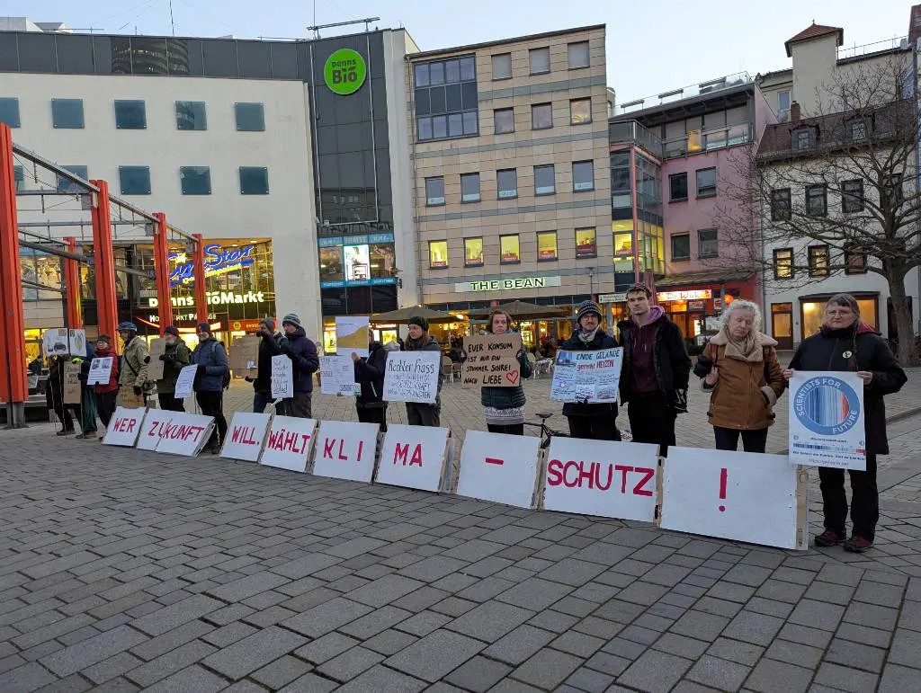 Eine Standdemo mit Transparenten zu verschiedenen Aspekten des Klimawandels und der Auswirkungen/ Beziehung zur Gesellschaft. Am Boden/ an den Beinen Tafeln mit dem Slogan: Wer Zukunft will - wählt Klimaschutz