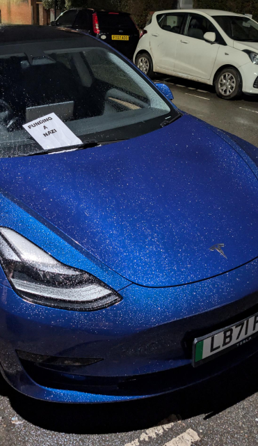 Photo of a parked Tesla with a sheet of paper on the windscreen, the words on the paper say "funding a nazi"