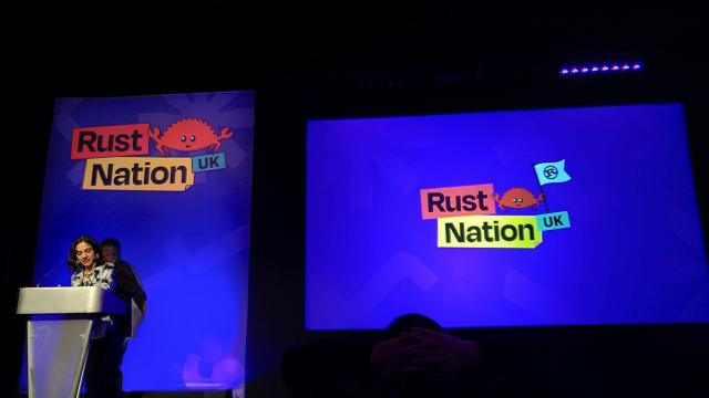 A photo of a conference stage with the RustNation UK visuals on a screen and banners.