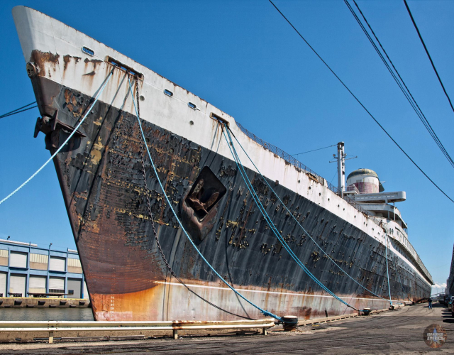 The prow of a black and white liner juts into the upper left portion of the frame like a knife. Covered in rust and moored by blue ropes, the other end of the liner seems impossibly far away.
