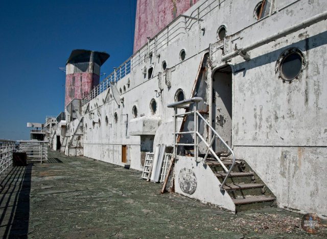 View down the deck of a large ocean liner, with a white wall dotted with portholes to the right. The towering red, white, and black funnels are visible above the wall.