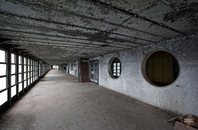 A long, barren stretch that was once the SS United States' promenade. Windows line the left wall and large circular holes are in the wall to the right next to a door.