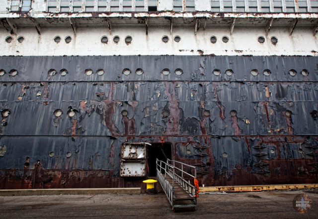 View of the side of a gargantuan liner covered with black and white peeling paint. A gangplank extends from the side to the road below.