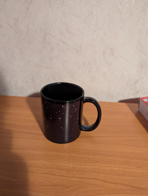 Das Bild zeigt eine schwarze Tasse mit weißen Sternen darauf, die auf einem braunen Tisch steht. Die Tasse hat einen weißen Henkel. Hinter der Tasse sieht man eine beigefarbene Wand.