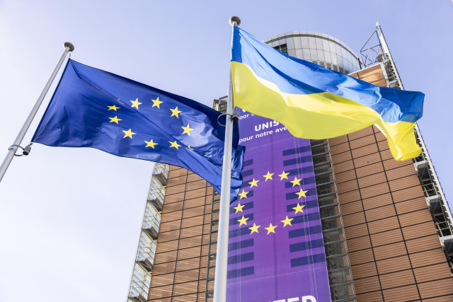 The EU flag and the Ukrainian flag are raised side by side. Behind them is the main building of the European Commission, the Berlaymont. There is an EU flag on the building. The sky is blue. 
