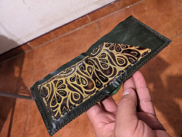 La cartera desplegada, luciendo el monedero que deja entrever una moneda de 10 pesotes. También se aprecian las costuras inferiores (tipo mocazin) y las laterales (puntada simple). El monedero es un bolsillo trapesoidal que se cierra cuando la cartera está cerrada por simple pliegue.