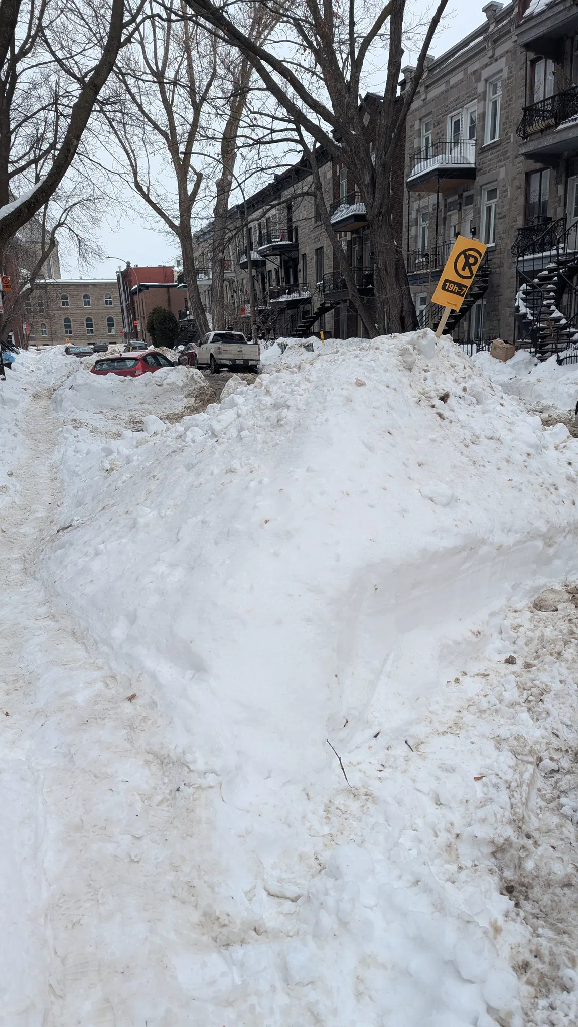 A street in Montreal. There is a lot of snow. In the snowbank is a temporary no parking sign. Slightly further up the road you can see the snow bank goes to the roof of a car that's been dig out.