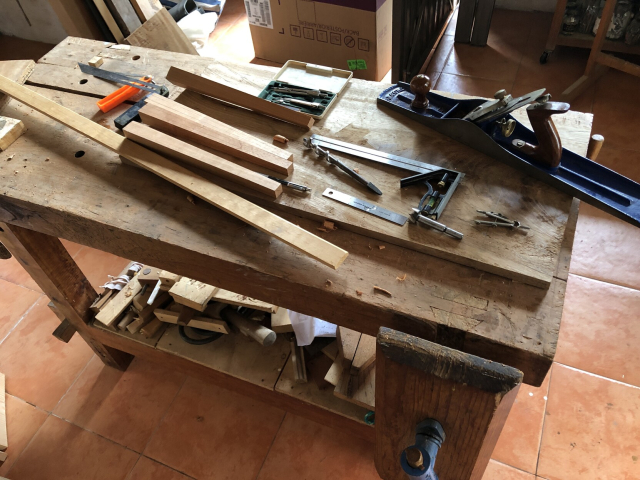 Banco de trabajo de carpintería cubierto de cosas: tabla para la superficie de la mesa, cuatro patas a medio hacer, una regla de madera, escuadras falsas y de combinación, compás, garlopa.
