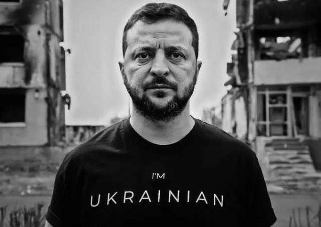 b&W photo of a man with close-cropped hair & beard (Zelenskyy) looking straight at us with an intense, serious gaze, wearing a black t-shirt with "I'm Ukrainian" written in white across the front, standing against a backdrop of bombed-out buildings, presumably in Ukraine.