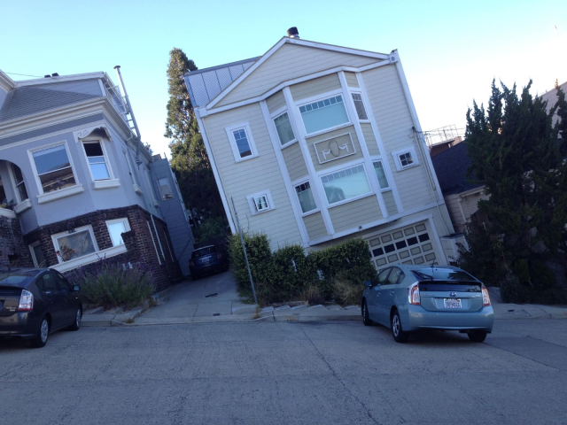 Two houses and cars on a steeply sloped street, creating an optical illusion of tilted buildings.