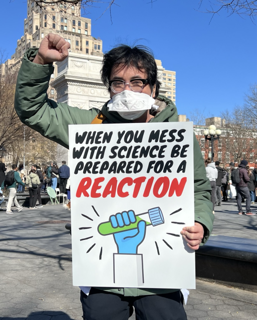 Dr. Lucky Tran at the NYC Stand Up For Science rally wearing at mask and holding a sign that says “When you mess with science, be prepared for a reaction!”