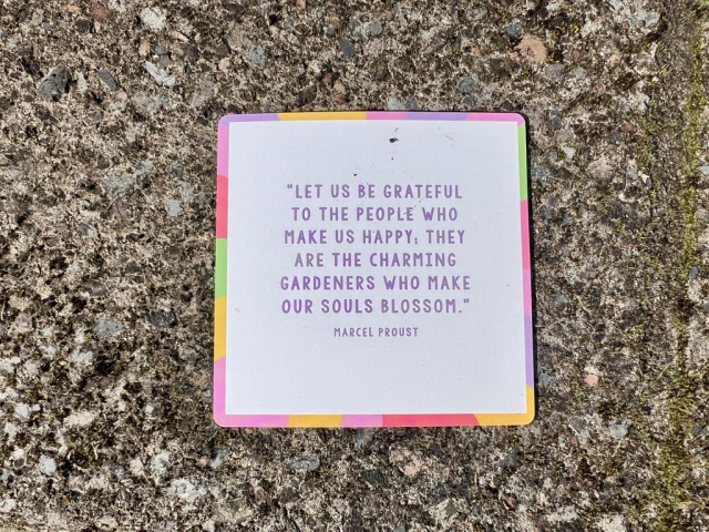 A 3 inch square card, with rainbow edging, that says, LET US BE GRATEFUL TO THE PEOPLE WHO MAKE US HAPPY, THEY ARE THE CHARMING GARDENERS WHO MAKE OUR SOULS BLOSSOM. - MARCEL PROUST