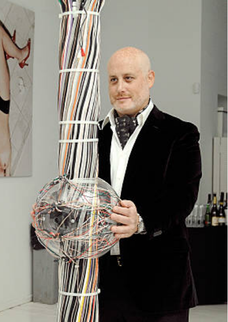 Bald middle aged man with a smirk wearing a suite and a cravat. He us clutching some kind of crystal ball with wires in it.