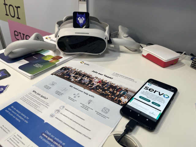 Leaflet showing hiring information about why and how to join our team at Igalia, resting on a table next to several devices like a Raspberry Pi, a mobile phone running Servo and a set of virtual reality glasses with a Wolvic sticker.