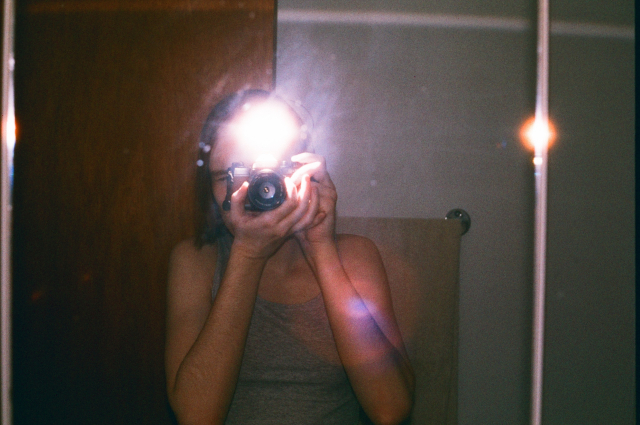 mirror selfie of me in a grey tank top holding a yashica fx-3 with the light of the flash covering my forehead