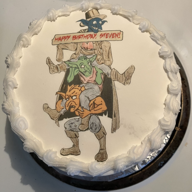 A round cake with white frosting and decorative swirls along the edge. In the center, there's an illustration of three goblins stacked on top of each other, all wearing a trench coat. The top character is also wearing a fedora. The middle one is green, and the bottom one is orange. There's a banner above them that reads "HAPPY BIRTHDAY, STEVEN!" in red letters.