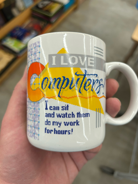 A late 80’s era Hallmark coffee mug with blocky, intentionally pixelated letters with a giant yellow triangle and squiggly line in the background. The text reads “I love computers! I can sit and watch them do my work for hours!”