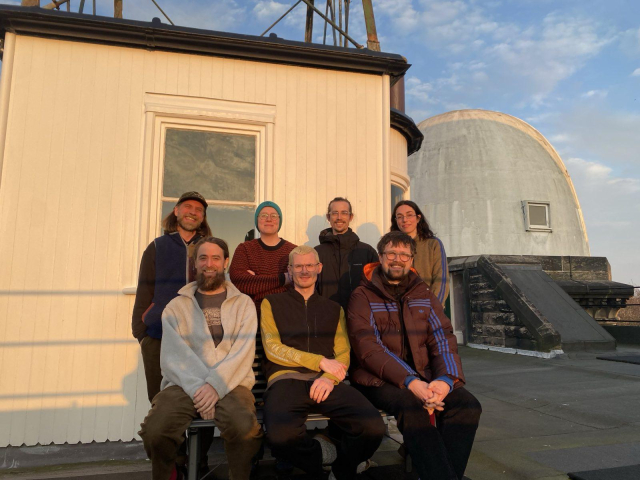 A group of seven people standing in front of a building, the sun is already low, in the background one can see the sky and another part of the building, an observation tower